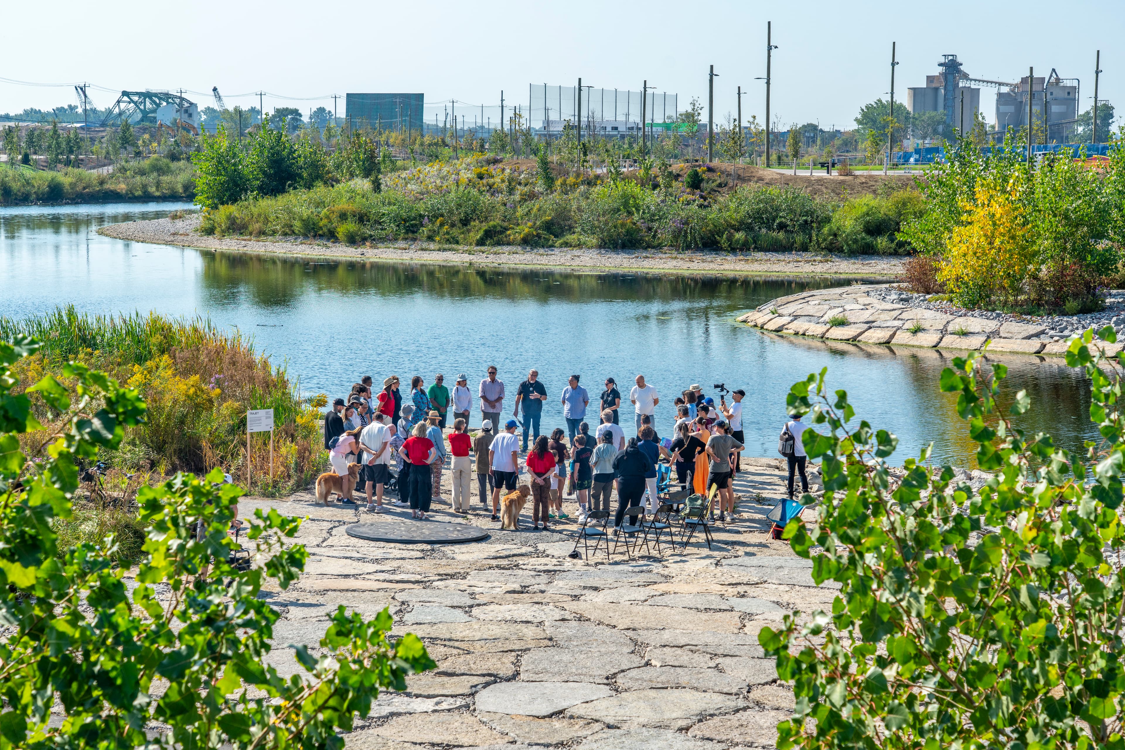A group of people standing beside Trajet, a circular sculpture with foot imprints. Green and yellow foliage surrounds the outdoor area.