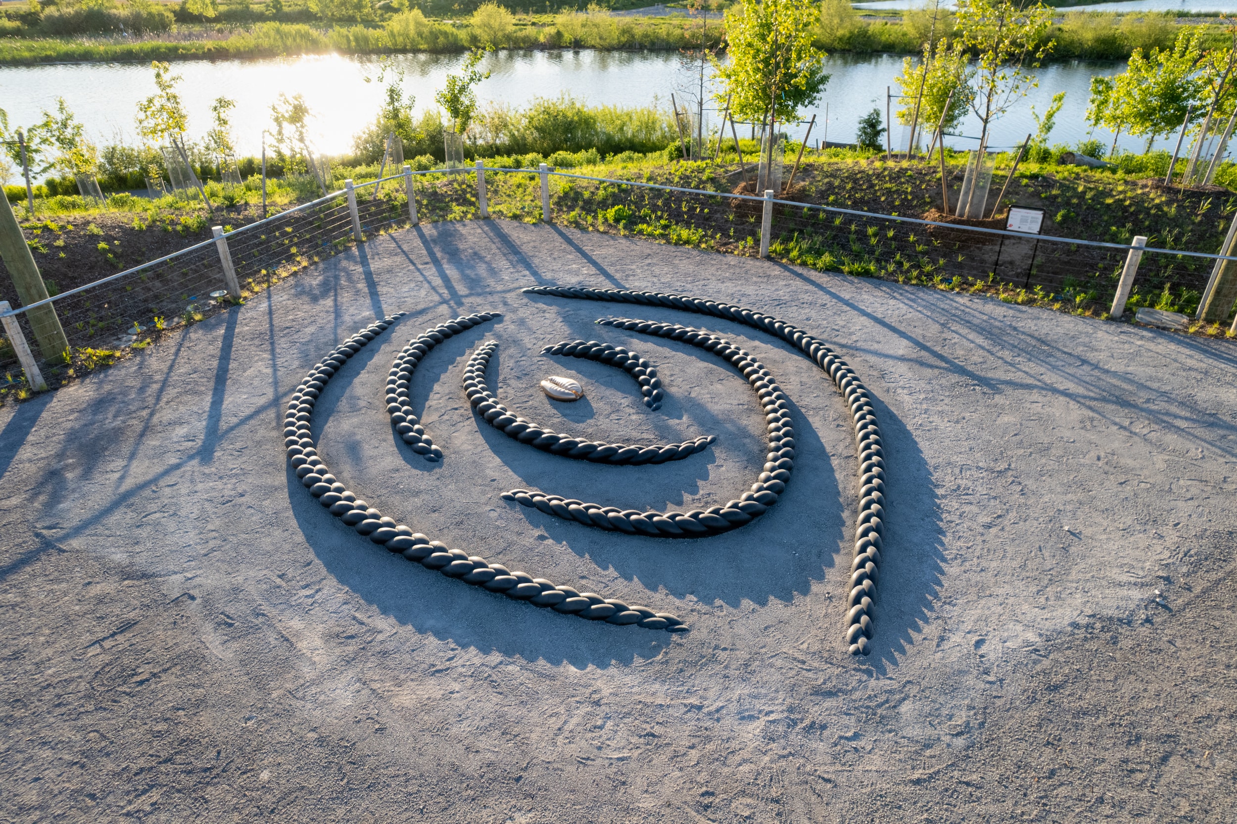 A large outdoor sculpture, Crown Act by Oluseye, features dark, thick, braid-like segments made of wood arranged in a spiral on a light gravel ground. A smaller, polished bronze cowrie shell sculpture rests in the centre of the spiral.