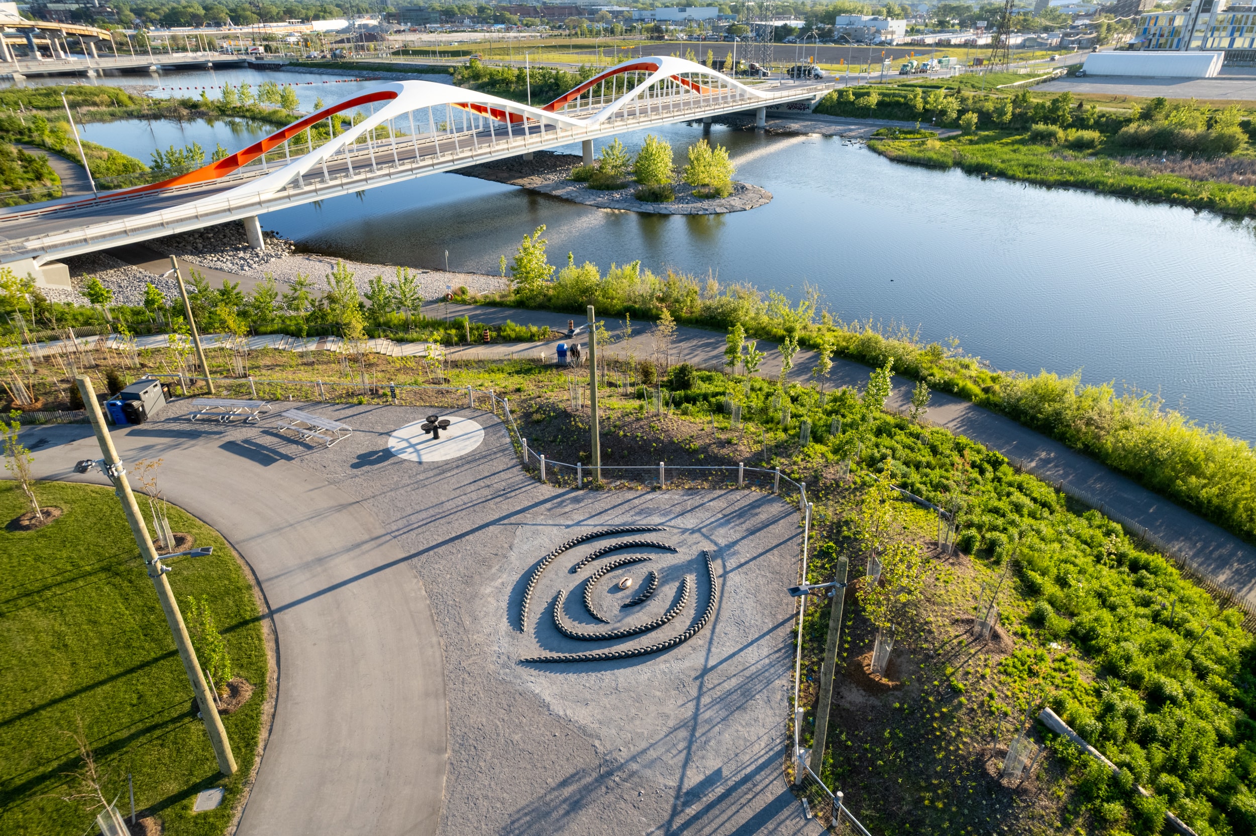 An aerial view shows Biidaasige Park with a white and red arch bridge spanning a dark blue river. Paved paths wind through green grass, featuring a large spiraling sculpture, Crown Act by Oluseye, made of dark stained wood and a circular target-like design. Buildings are visible in the distant background.