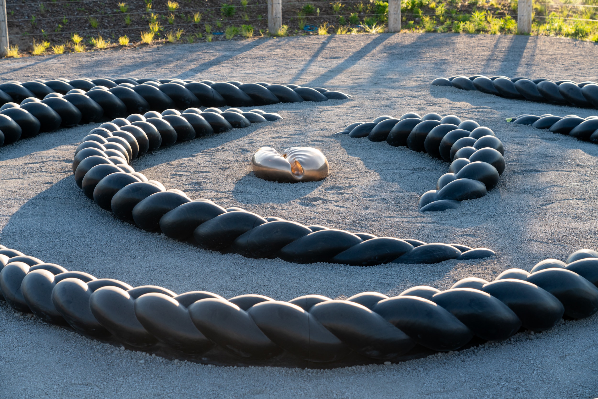A large outdoor sculpture, Crown Act by Oluseye, features dark, thick, braid-like segments made of wood arranged in a spiral on a light gravel ground. A smaller, polished bronze cowrie shell sculpture rests in the centre of the spiral.