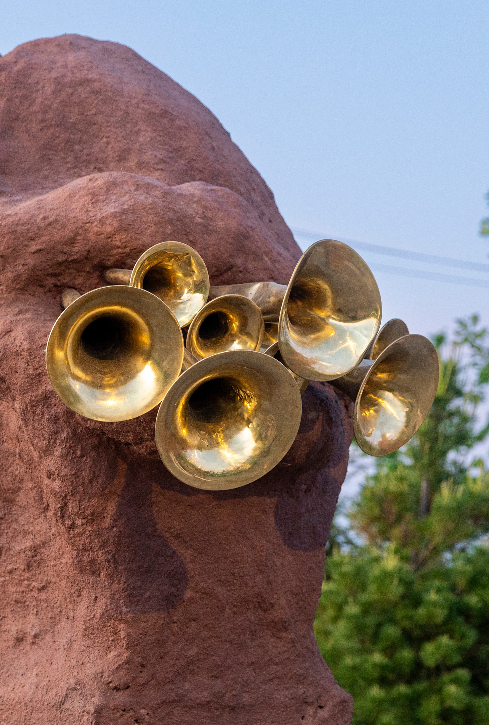 A cluster of golden brass instrument bells protrudes from a large, reddish-brown, textured sculpture.
