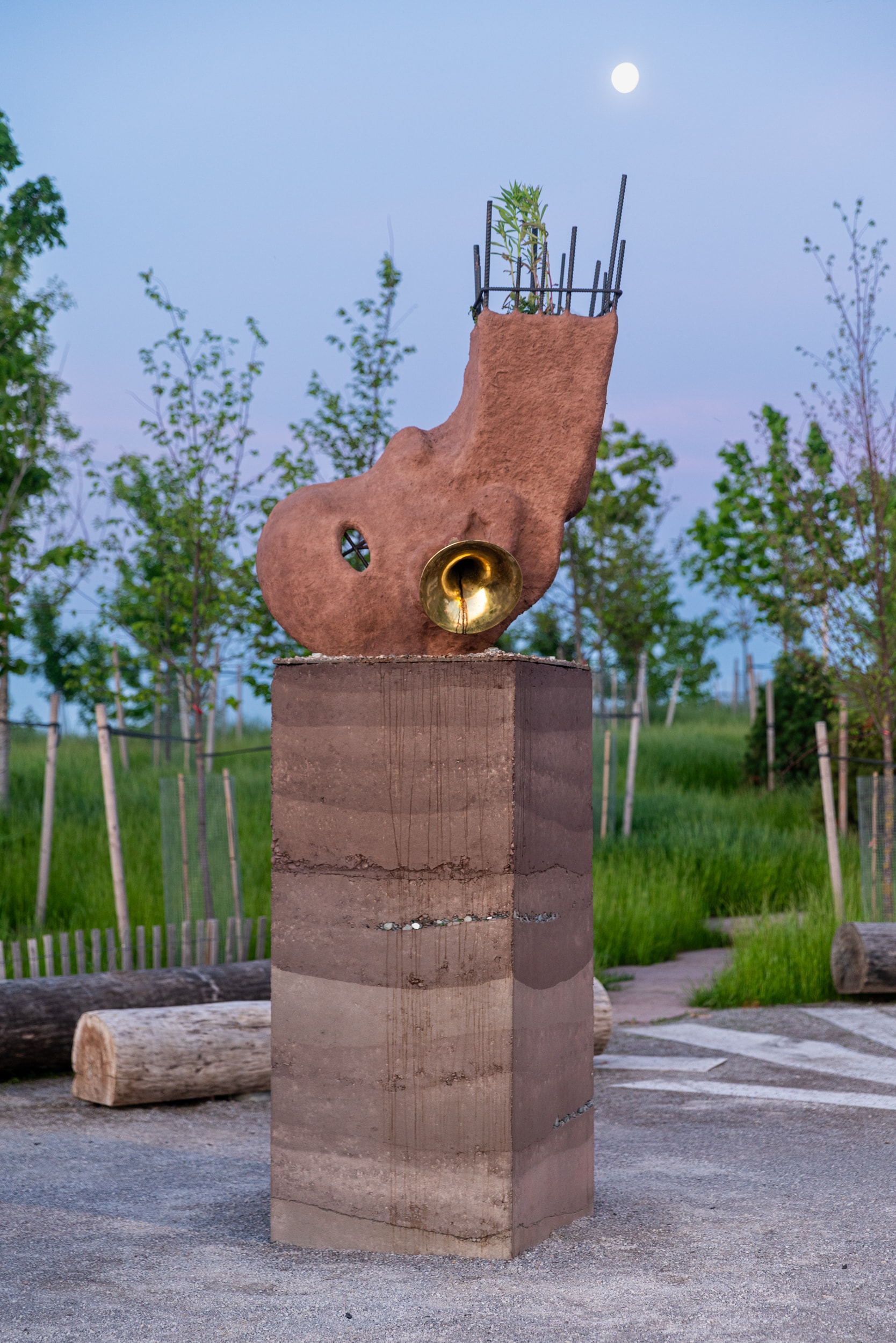 A reddish-brown, ear-shaped sculpture with a golden brass trumpet embedded in its side. It rests on a square, layered pedestal of similar earthy material, set on a gravel path with young trees in the background.