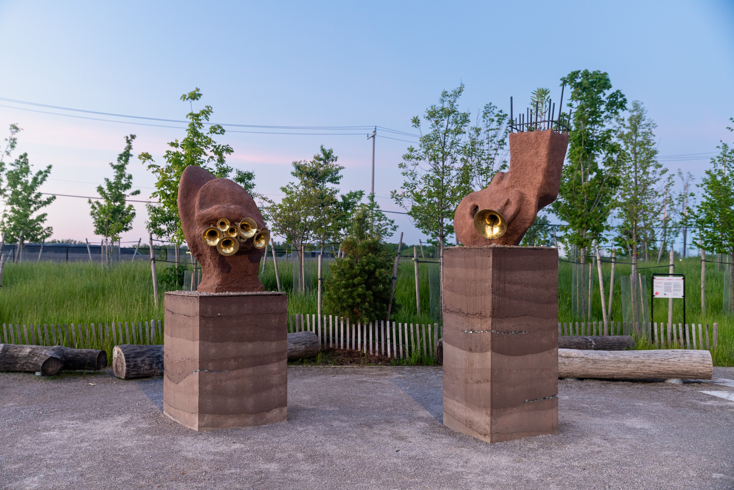 Two large, organic, reddish-brown sculptures made of cob stand on layered rammed earth pedestals on a gravel path. The left sculpture features multiple brass horn-like elements, and the right sculpture has similar brass elements with an irregular top revealing internal rebar. Young trees and grass are visible in the background.