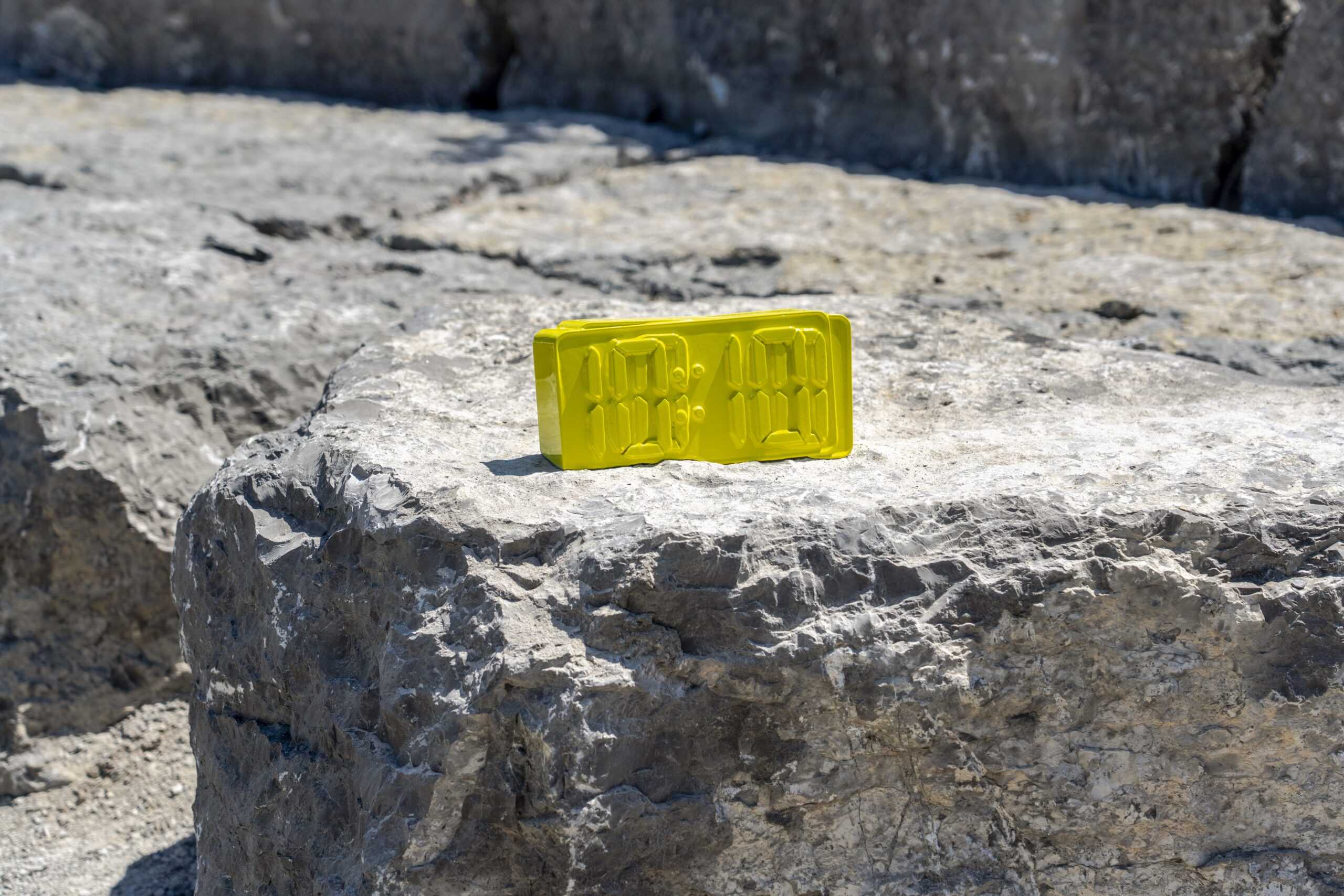 A bright yellow sculpture of a digital alarm clock rests on a large, textured grey rock.