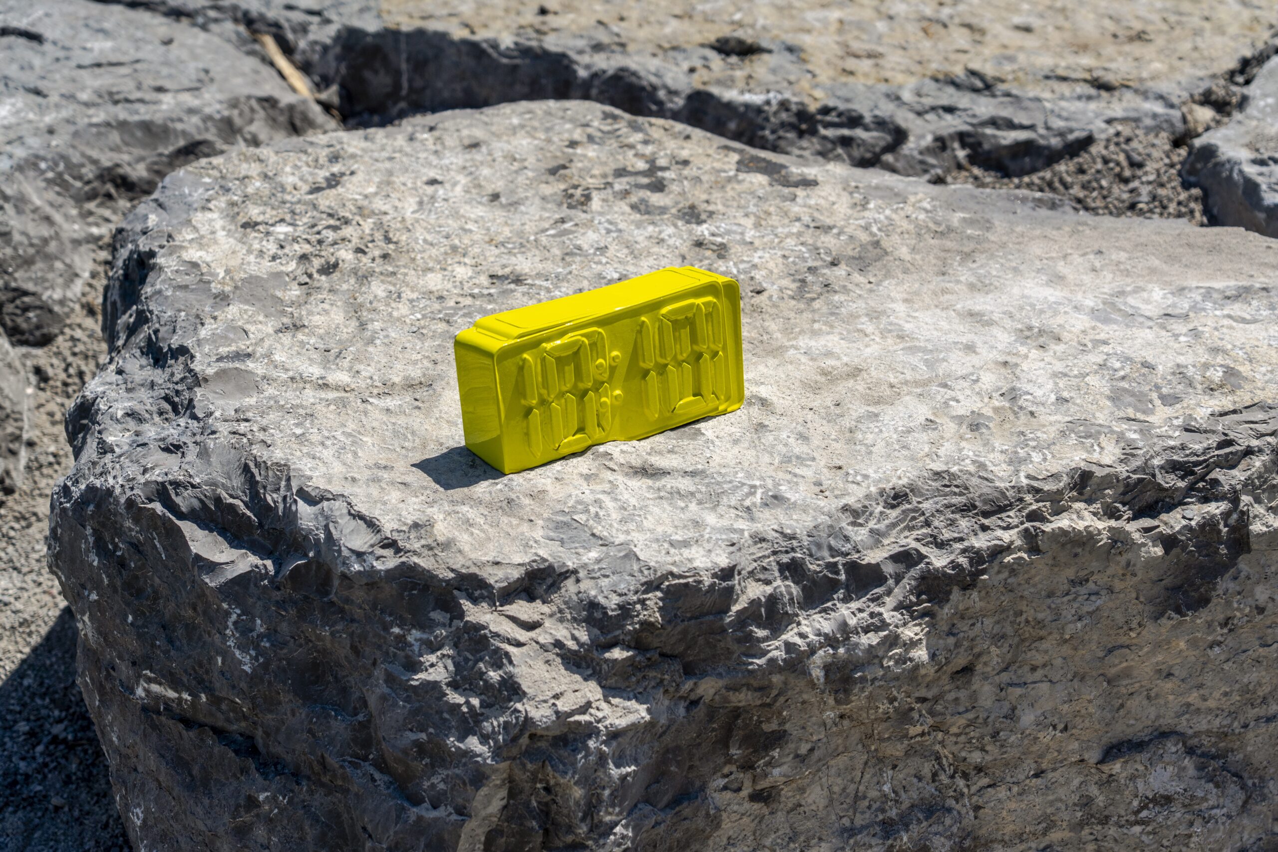 A bright yellow sculpture of a digital alarm clock rests on a large, textured grey rock.