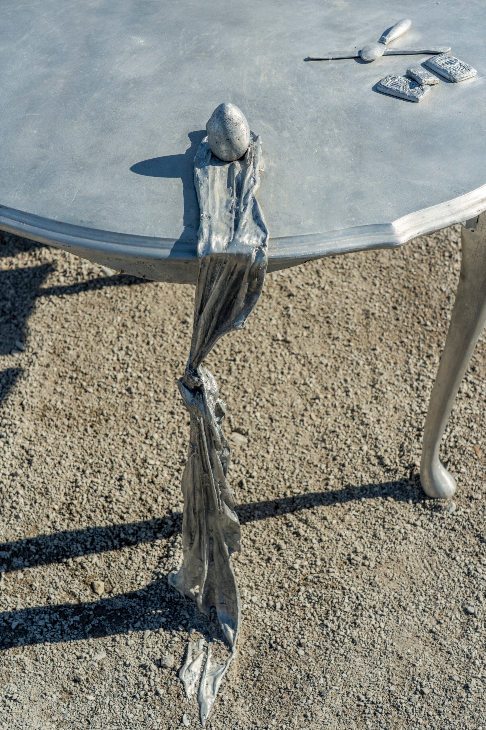 A silver-colored, octagonal table stands on a gravel surface. A draped, fabric-like element, also silver, hangs from the table's edge to the ground. On the table surface are a small silver sphere and several flat, rectangular silver objects.
