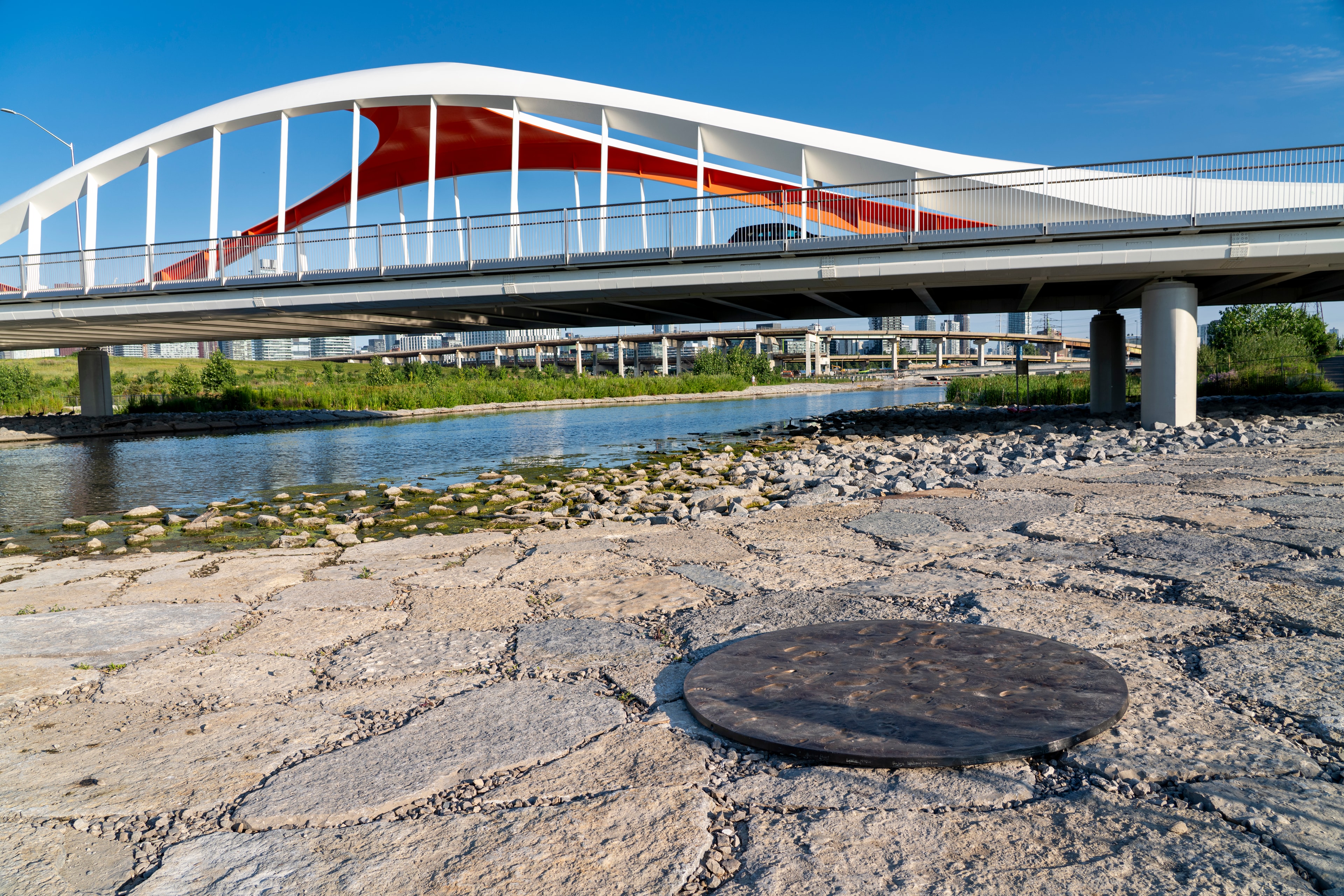 A modern white arched pedestrian bridge with red and orange fabric panels spans a river. In the foreground, a dark circular disc is embedded in a stone-paved bank. Distant city buildings are visible under a clear blue sky.