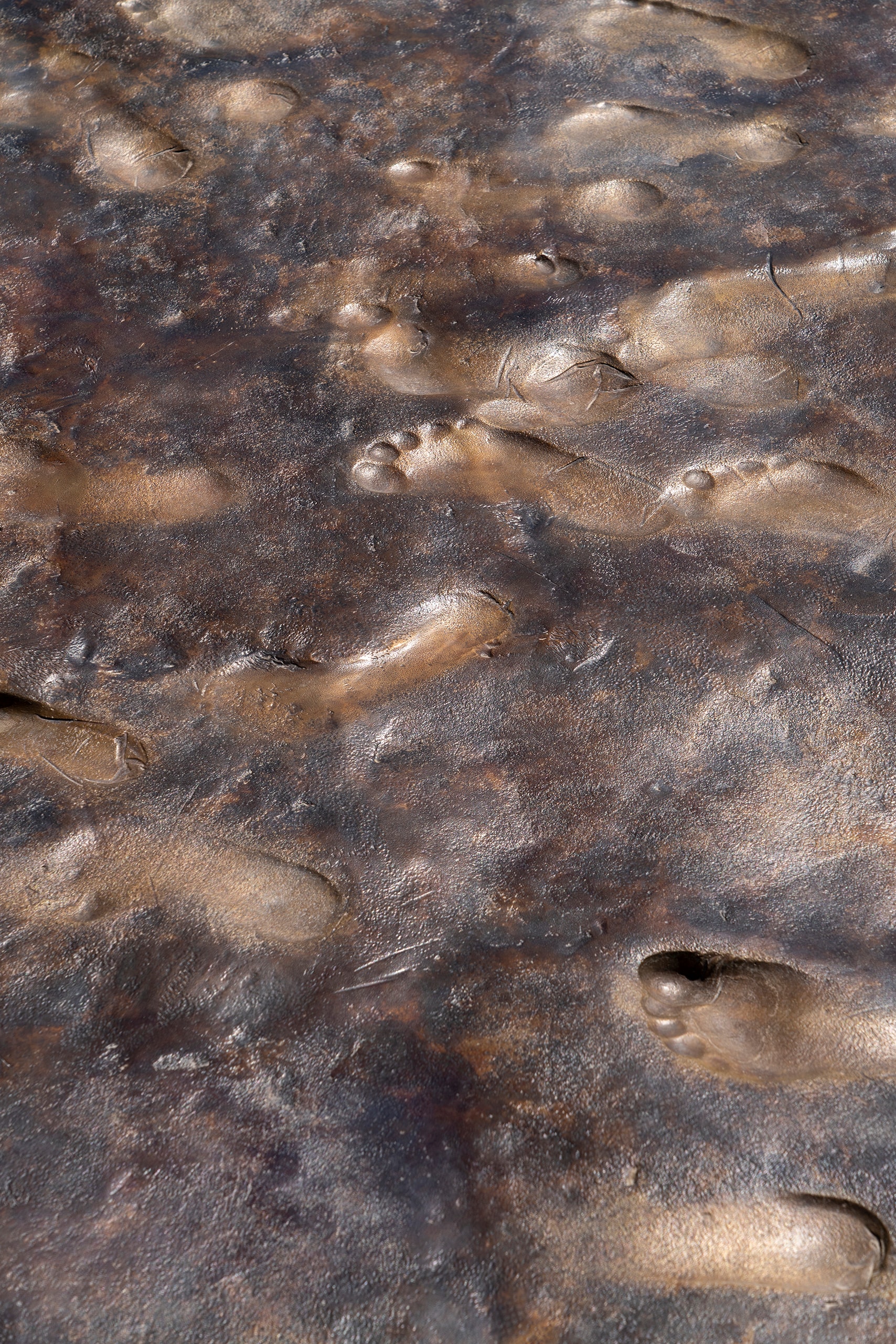 A dark, textured metallic surface is covered with numerous impressions of human footprints and handprints. The prints are recessed into the material, which has a bronze-like sheen.