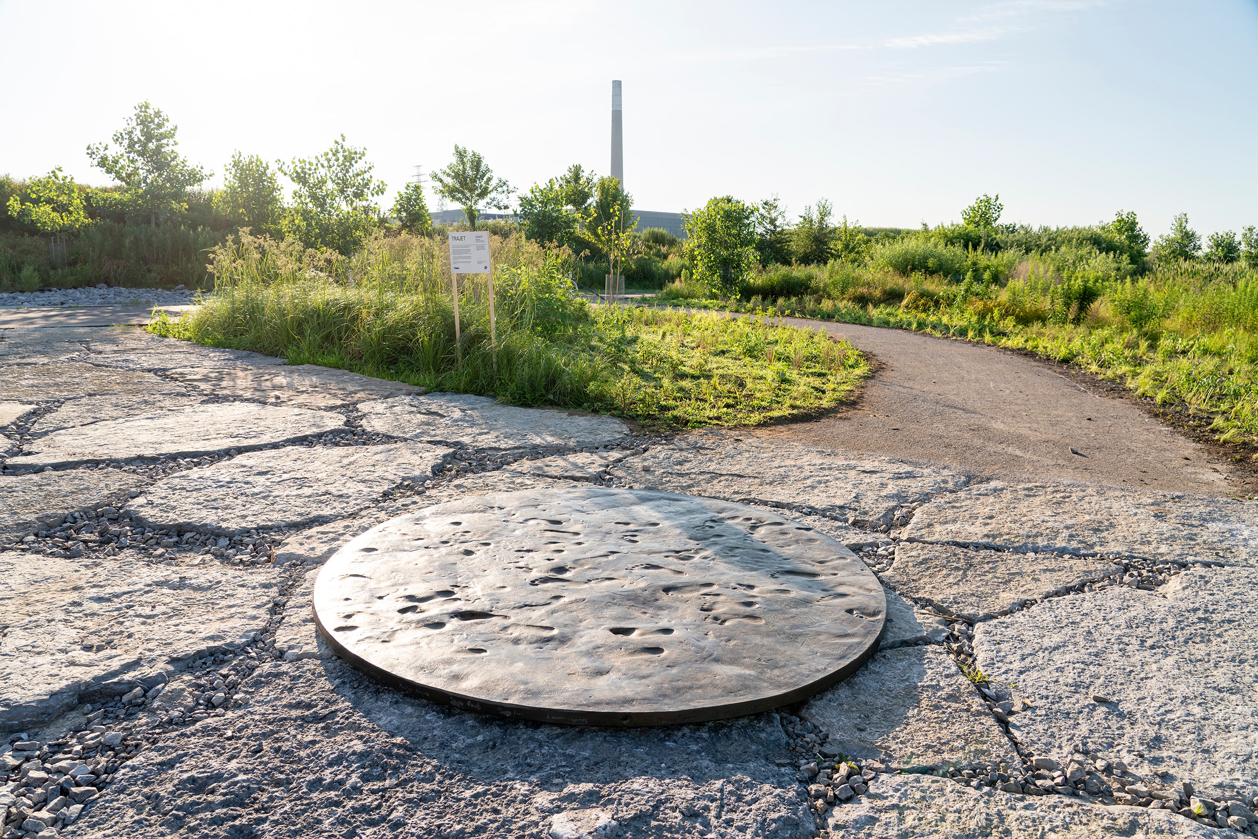A large, dark circular sculpture with footprints embedded in its surface is integrated in a path of irregular stone slabs. The path curves through a grassy landscape with trees, and a tall industrial chimney stands in the distance.