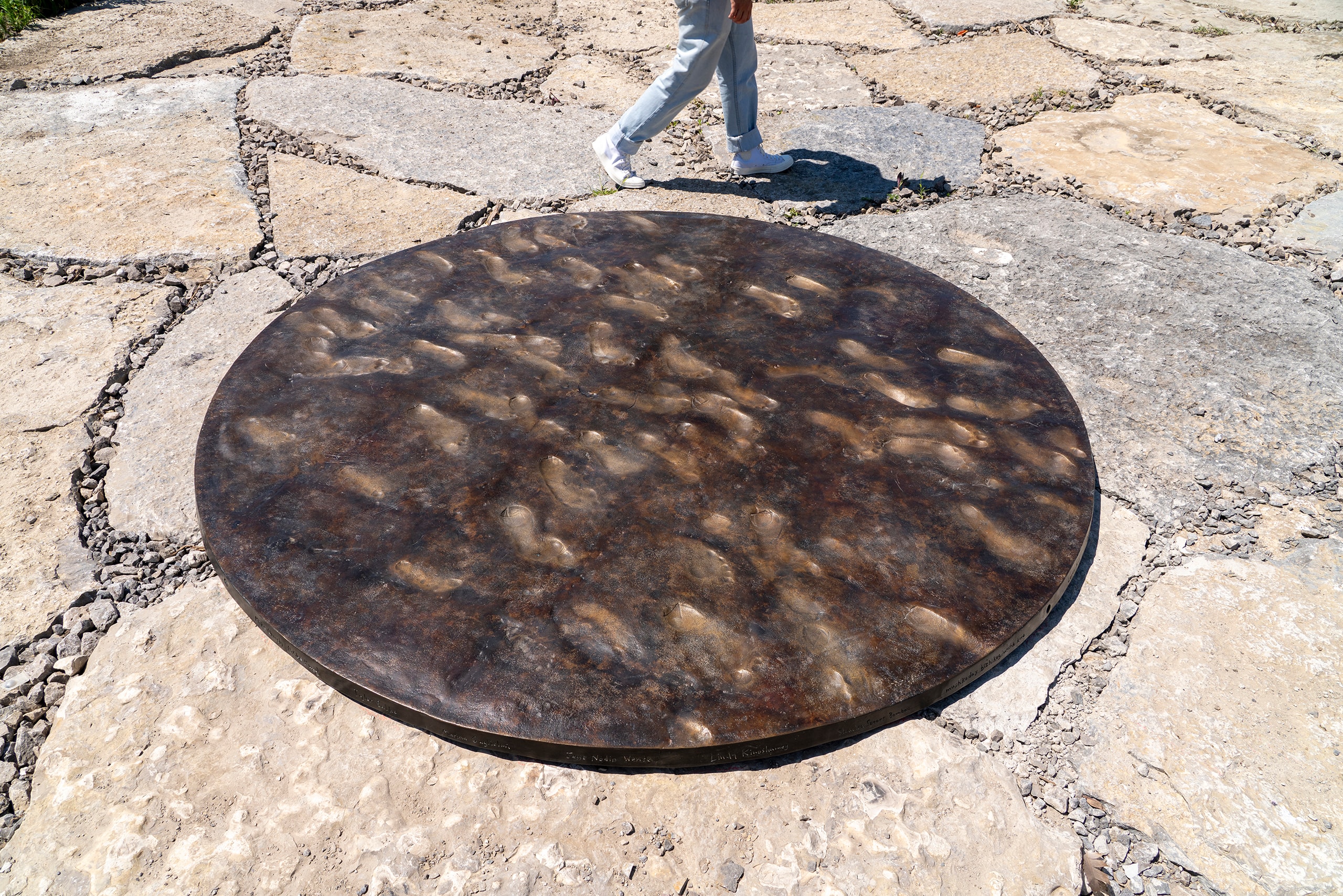 A large, dark circular sculpture, Trajet, with numerous footprint impressions, is set into a light-colored flagstone path. The lower legs of a person in light pants and white shoes walk past in the upper right.