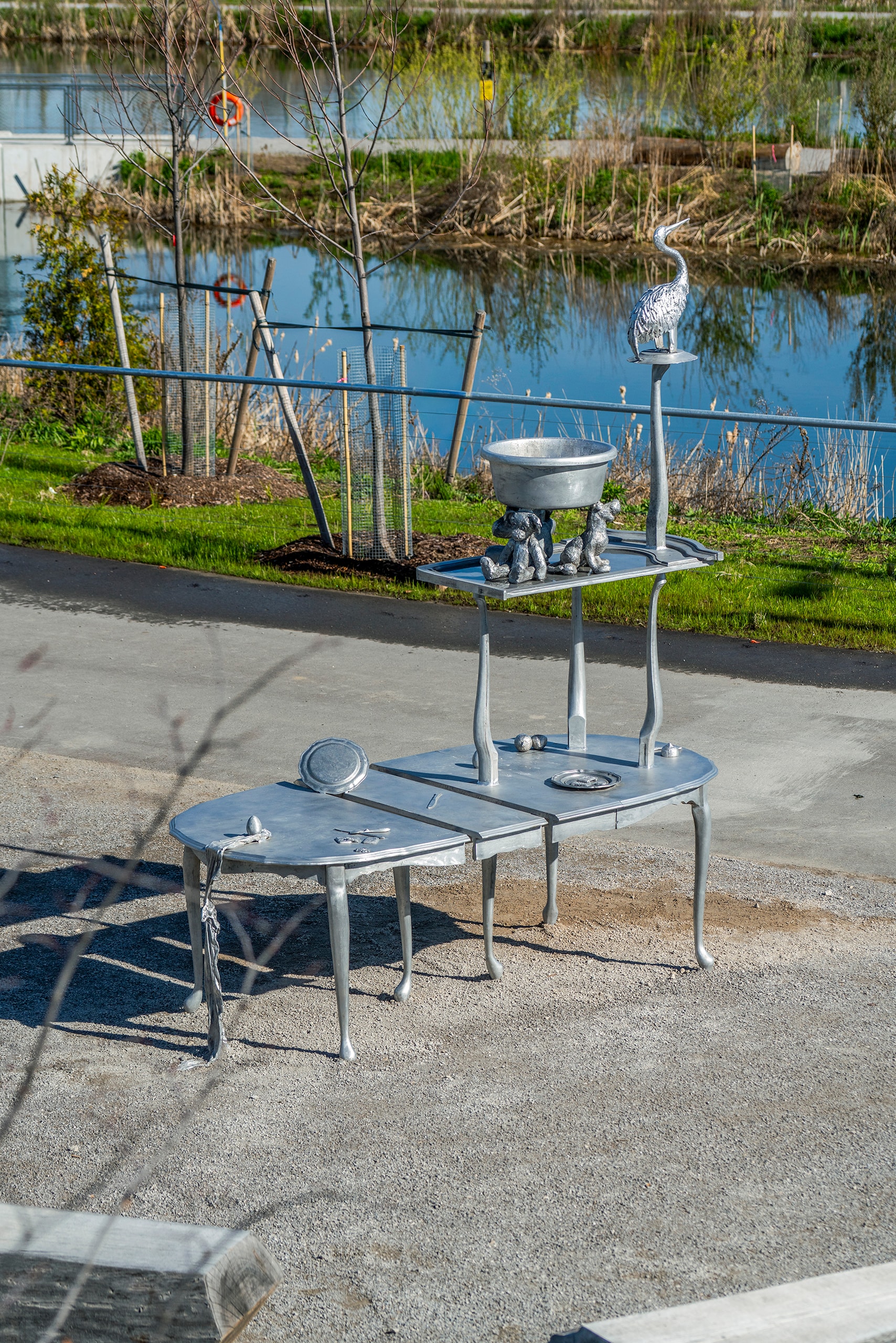 A silver-coloured metal sculpture, Homing by Nadia Belerique and Tony Romano, features two tables, one stacked above and slightly behind the other. The lower table holds a plate and cutlery. The upper table displays a large bowl, several small duckling figures, and a tall crane-like bird on a pedestal. The sculpture stands on a paved path next to a body of water with reeds.