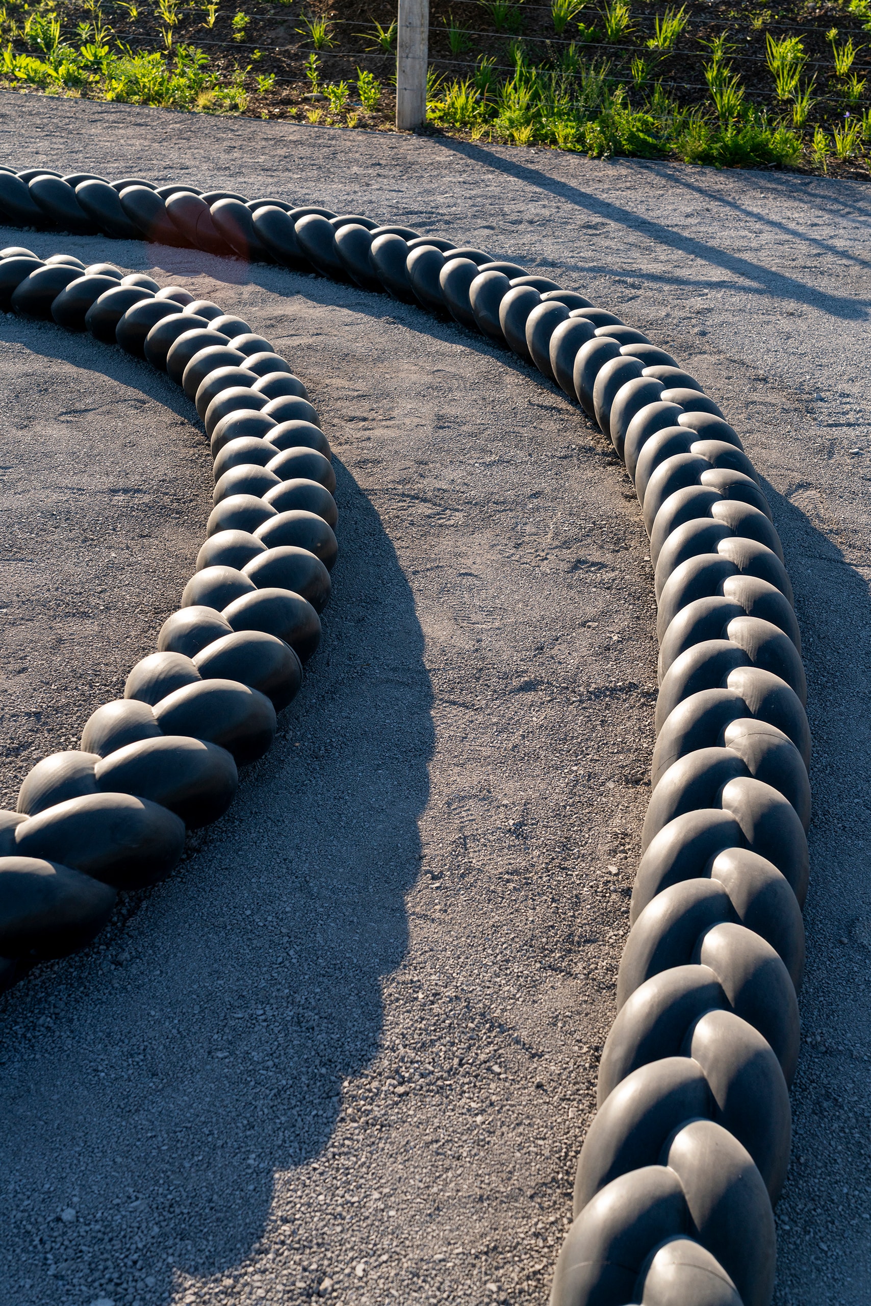 A large, dark, braided sculpture, Crown Act by Oluseye, winds across a light gravel path, casting shadows. The sculpture is composed of numerous rounded segments linked together.