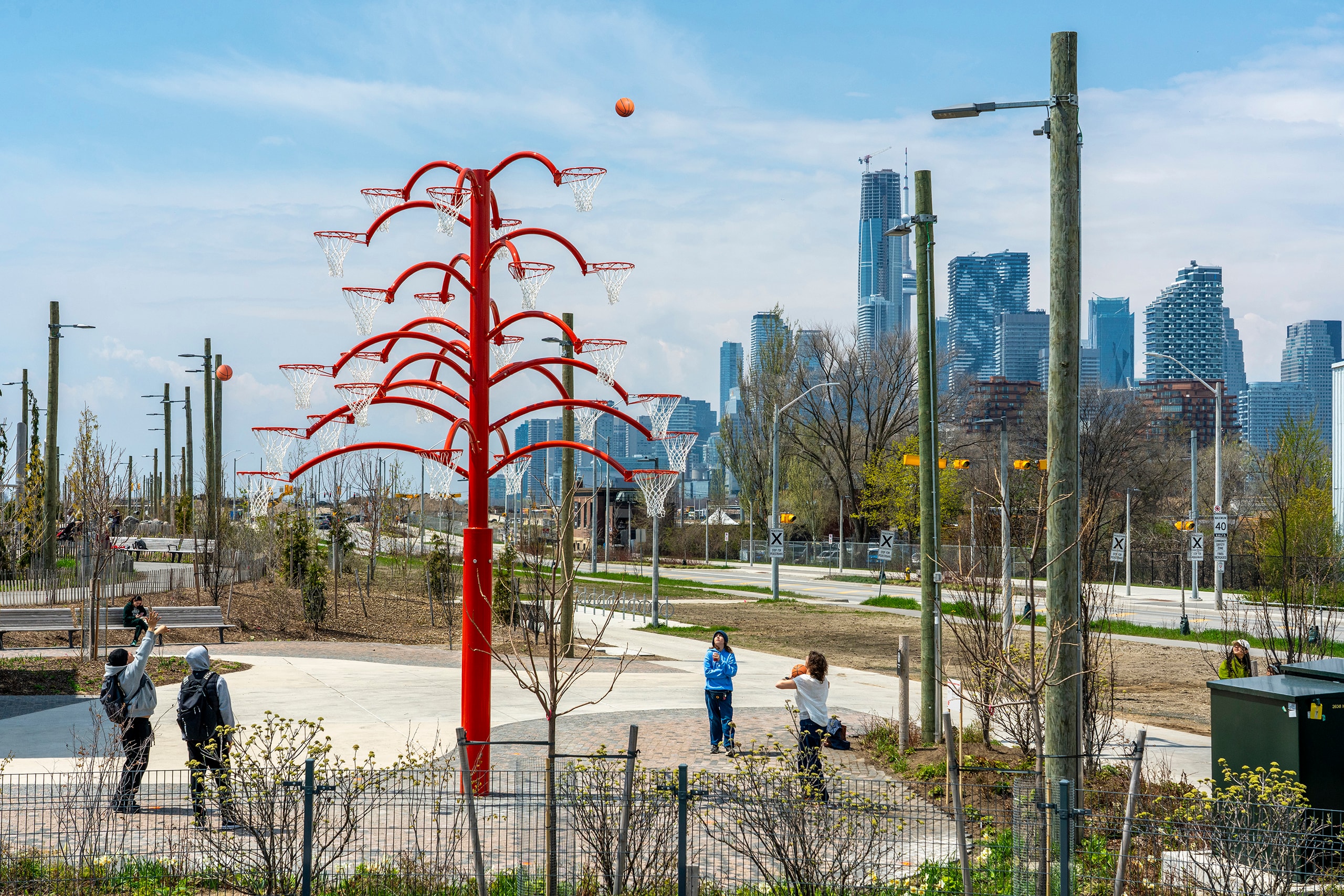A tall, bright red, tree-like sculpture with multiple basketball hoops stands in an urban park (Biidaasige). An orange basketball is mid-air near the top of the sculpture, while four people play below. A city skyline with modern buildings is visible in the background.