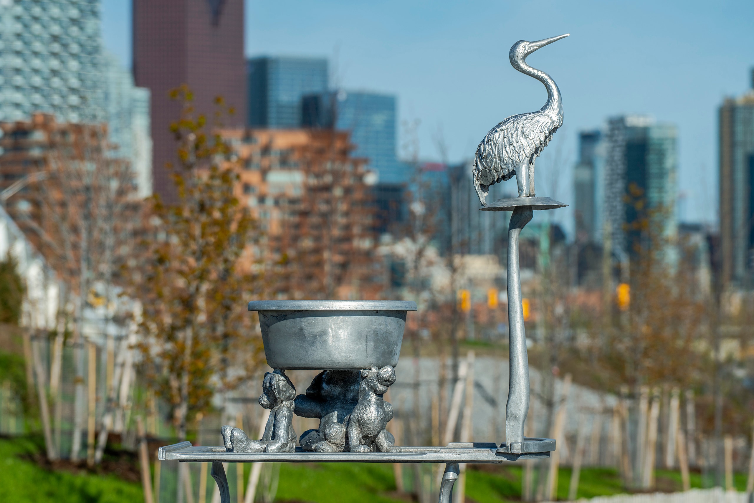 An aluminum metal sculpture features a heron standing on a tall post, next to a shallow basin supported by three small, aluminum animal figures. The outdoor sculpture is set against a blurred background of city buildings and trees.