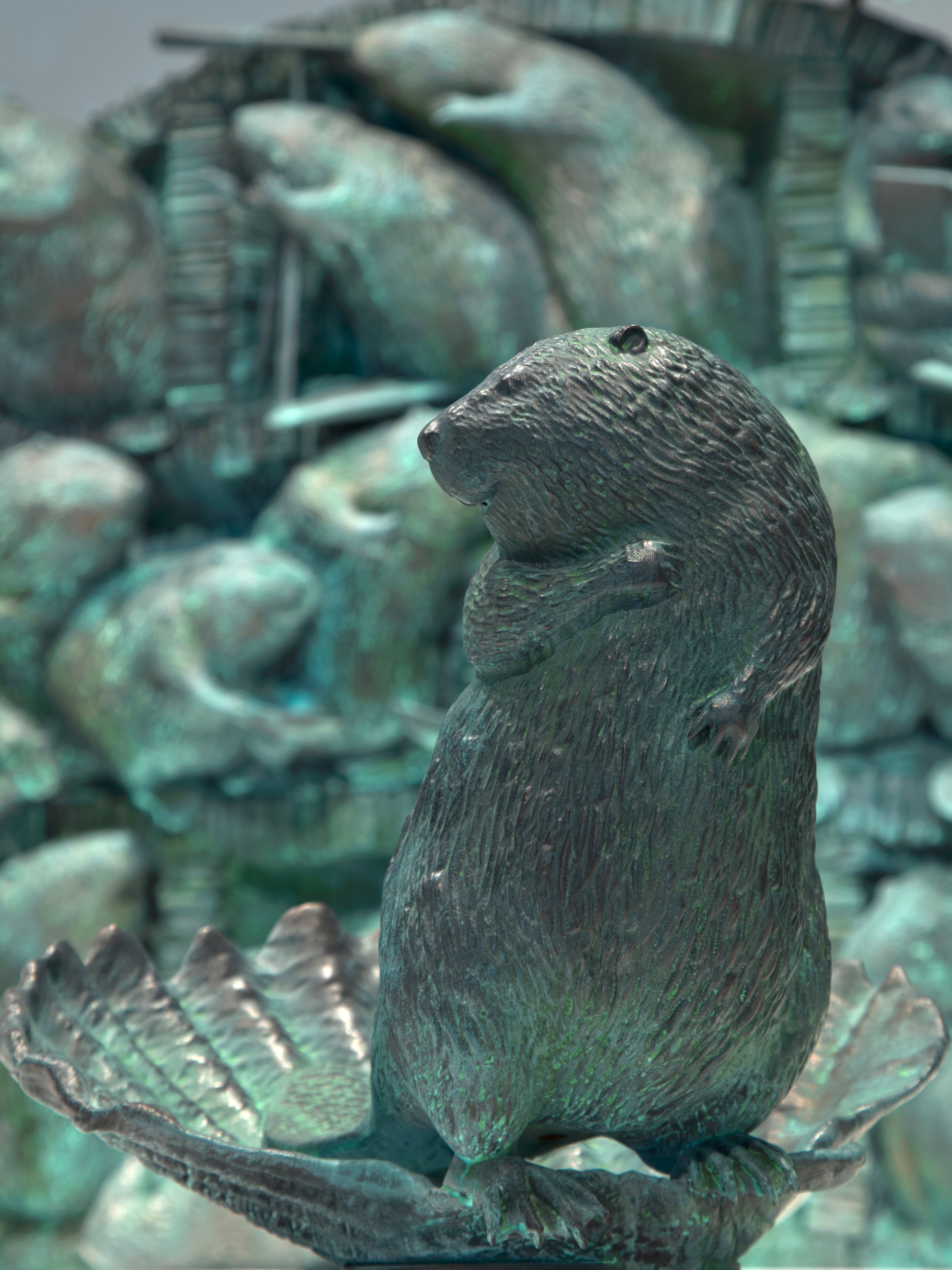A dark, greenish-bronze sculpture of a beaver standing upright on a shell-shaped base. The beaver has textured fur and its front paws are held to its chest. Other beaver sculptures are blurred in the background.