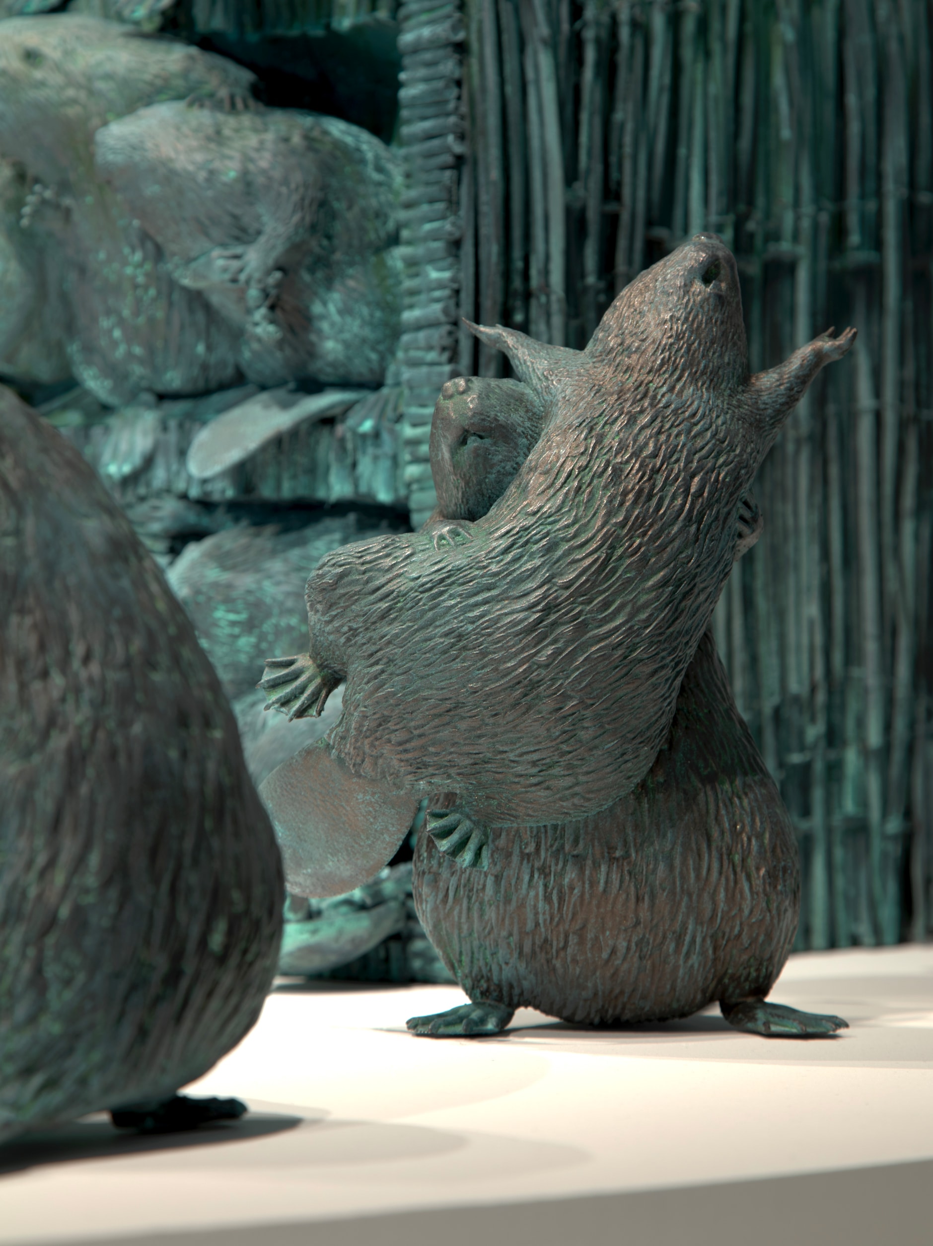A dark, greenish-bronze sculpture of a beaver dancing with another beaver. The beavers have textured fur. Other beaver sculptures are blurred in the background.