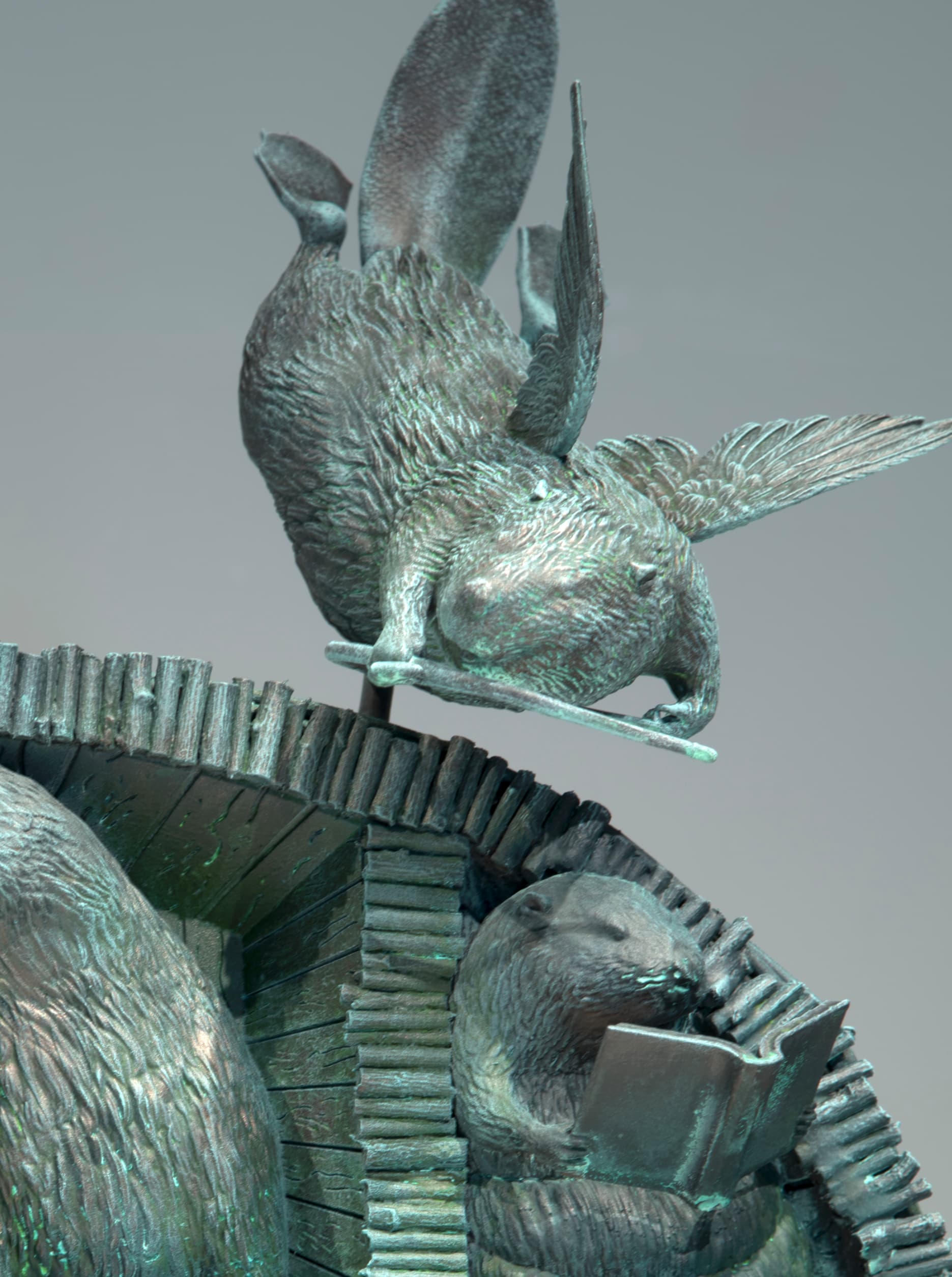 A bronze sculpture depicts a flying beaver perched on a beaver lodge, with a beaver below reading an open book. The sculpture has a greenish patina.