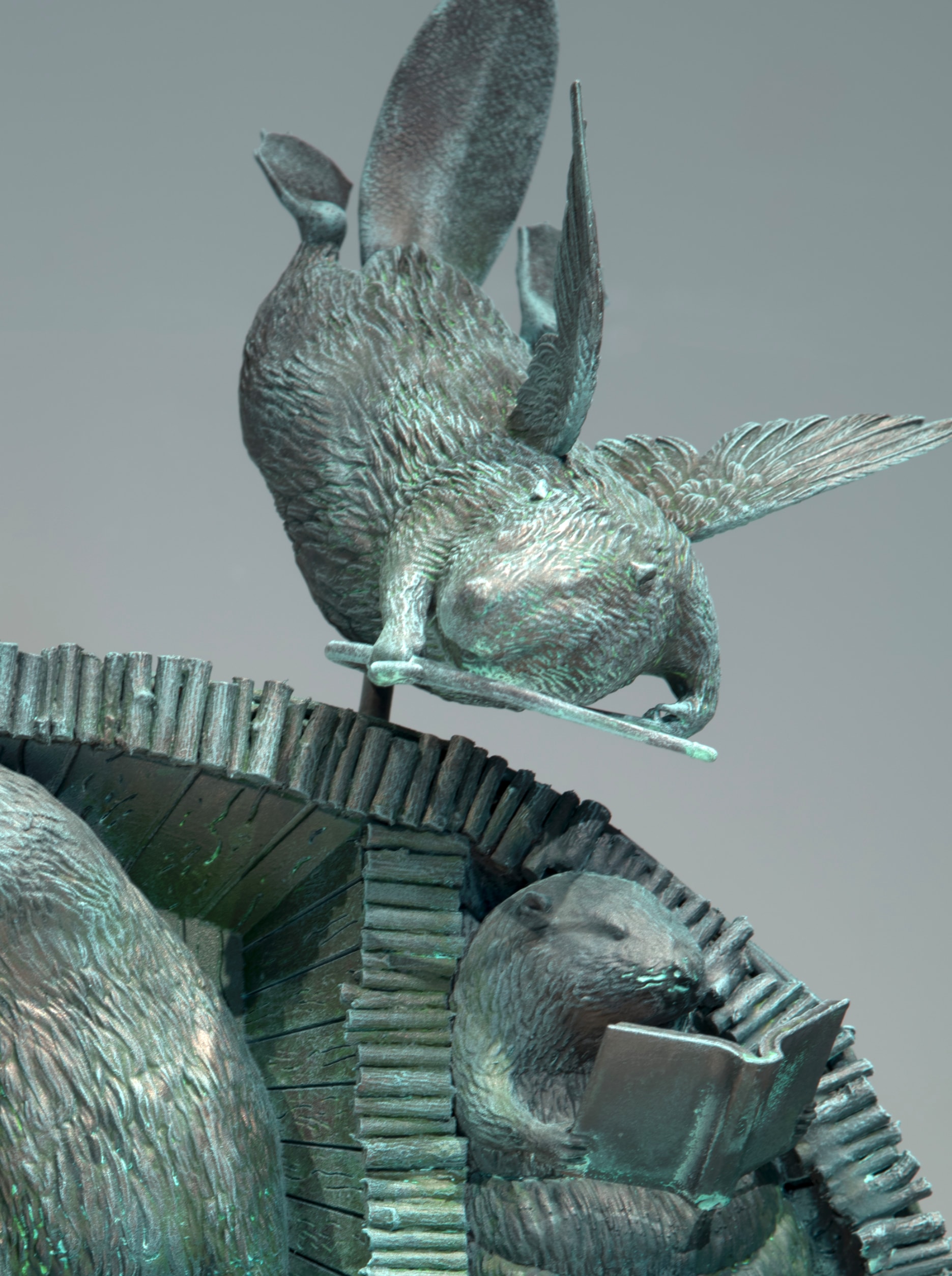 A bronze sculpture depicts a flying beaver perched on a beaver lodge, with a beaver below reading an open book. The sculpture has a greenish patina.