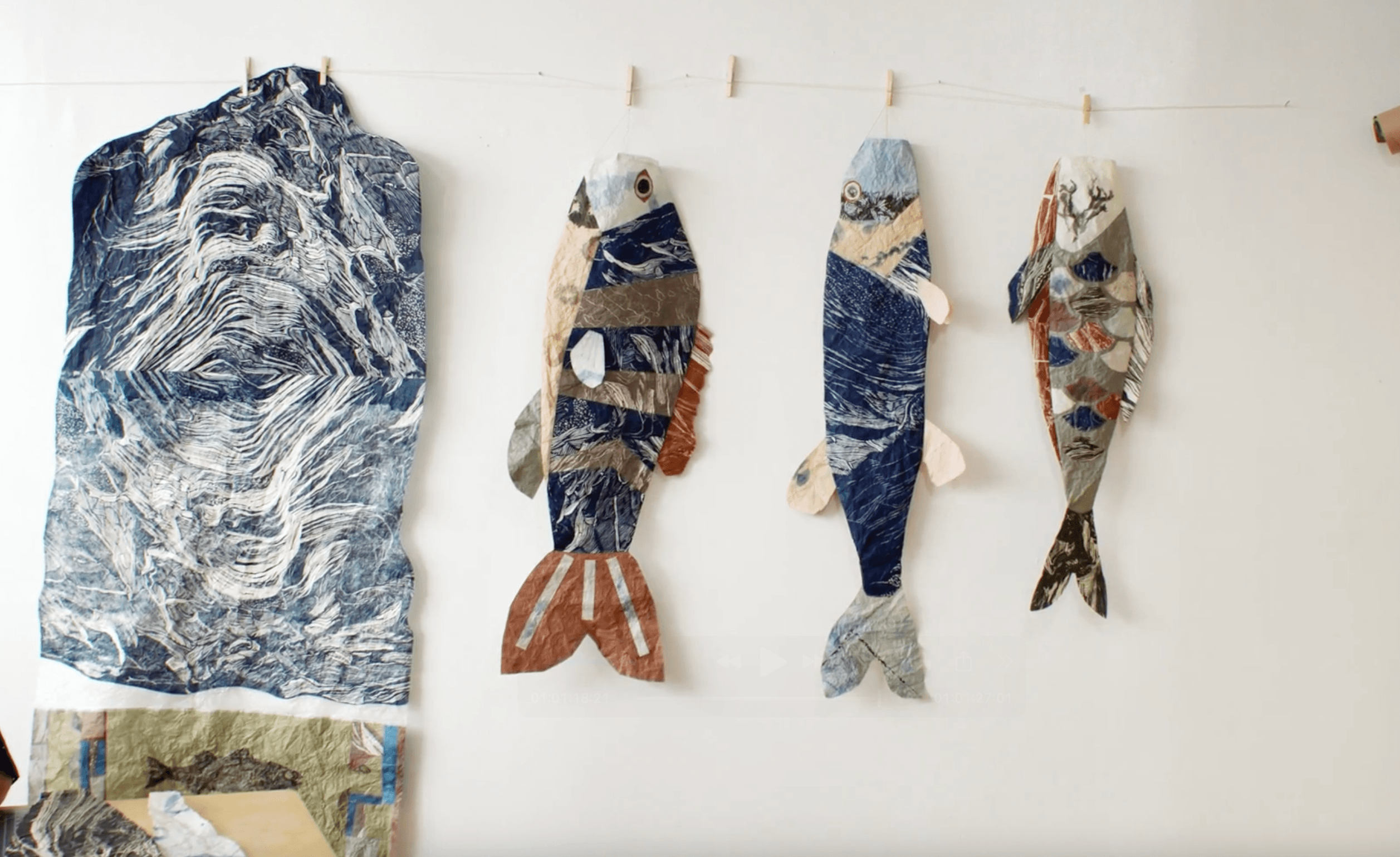 Several paper collage artworks hang against a white wall. On the left is a large rectangular piece with blue and white swirling patterns. To its right, three fish-shaped artworks, made from patterned paper in shades of blue, brown, and white, hang from a string with clothespins.