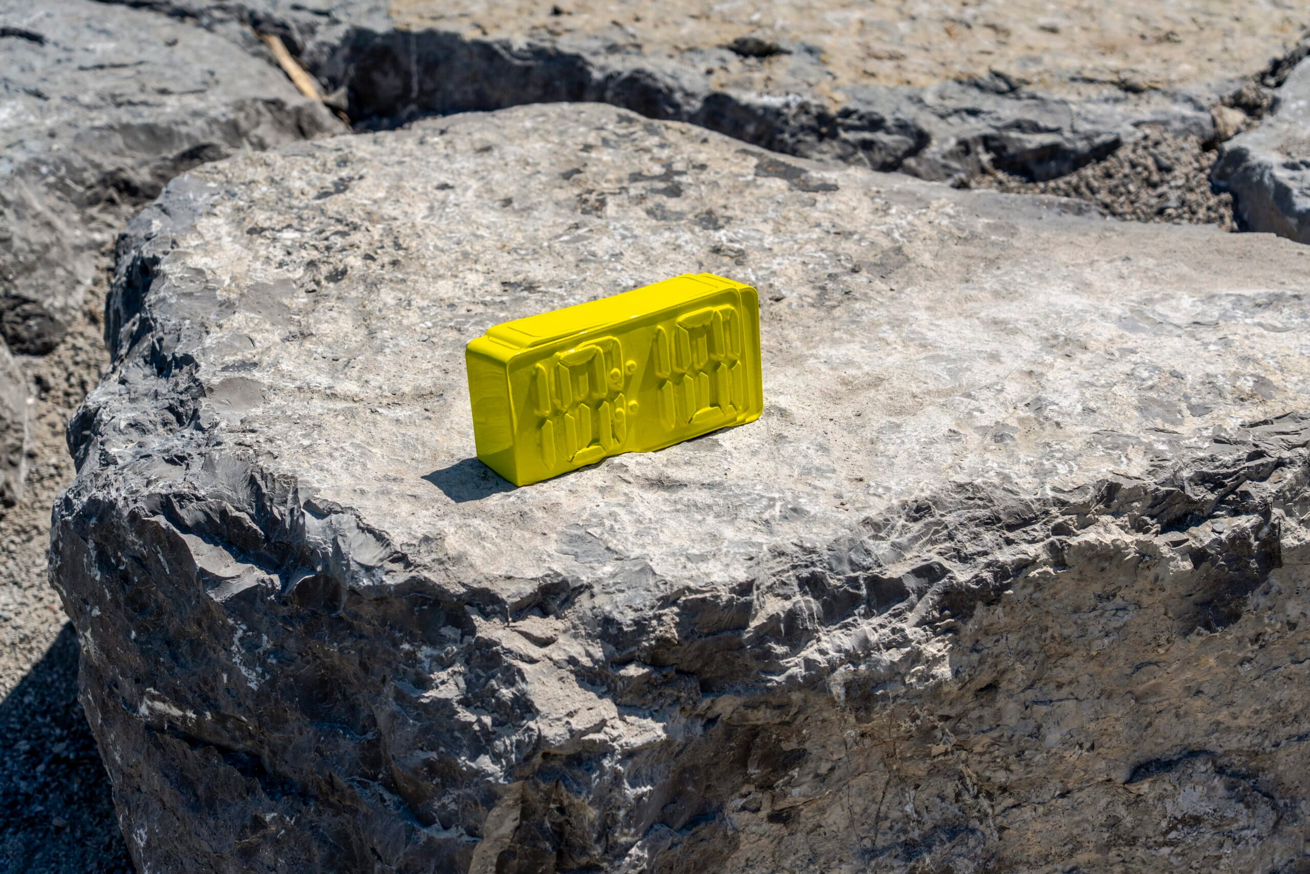 A bright yellow sculpture of a digital alarm clock rests on a large, textured grey rock.