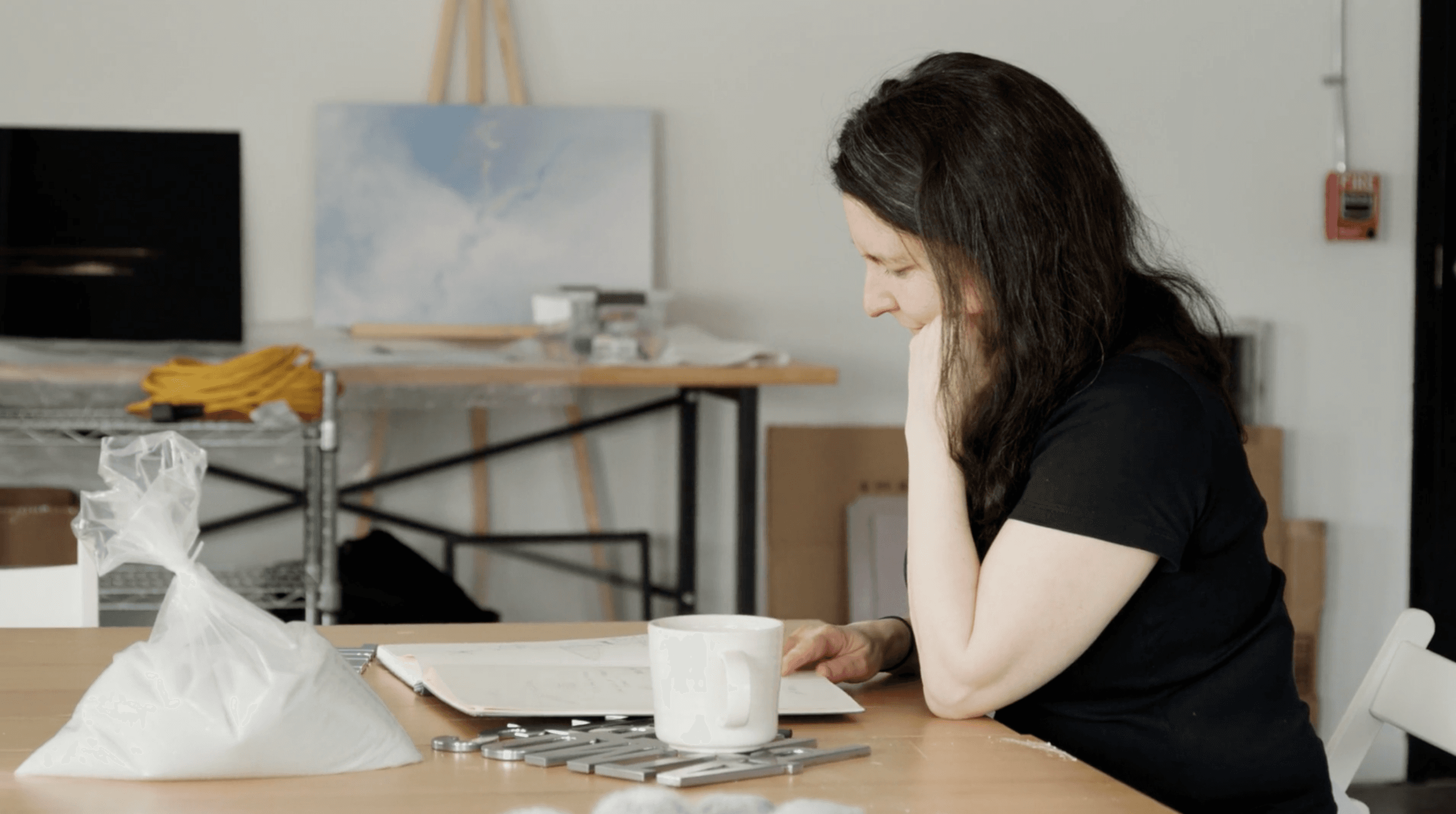 A woman with dark hair sits at a light wooden table, looking down at an open book. A white mug, metal tools, and a clear bag of white granular material are on the table. An art studio with a painting on an easel is visible in the background.