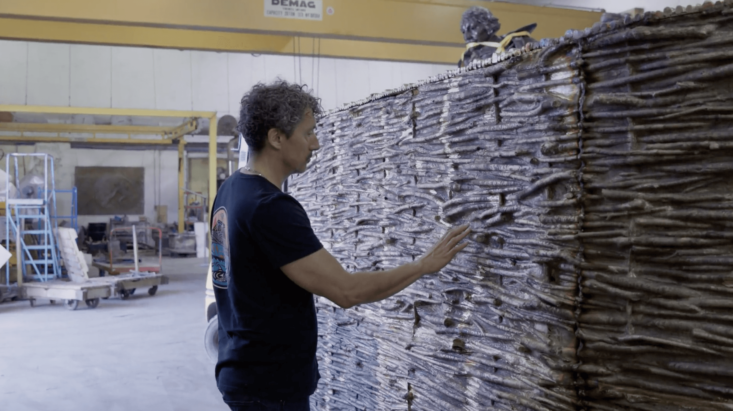 The artist Kent Monkman wearing a dark t-shirt touches a large, dark sculpture composed of numerous interwoven, branch-like elements, creating a rough texture. The sculpture stands in an industrial workshop with machinery visible in the background.