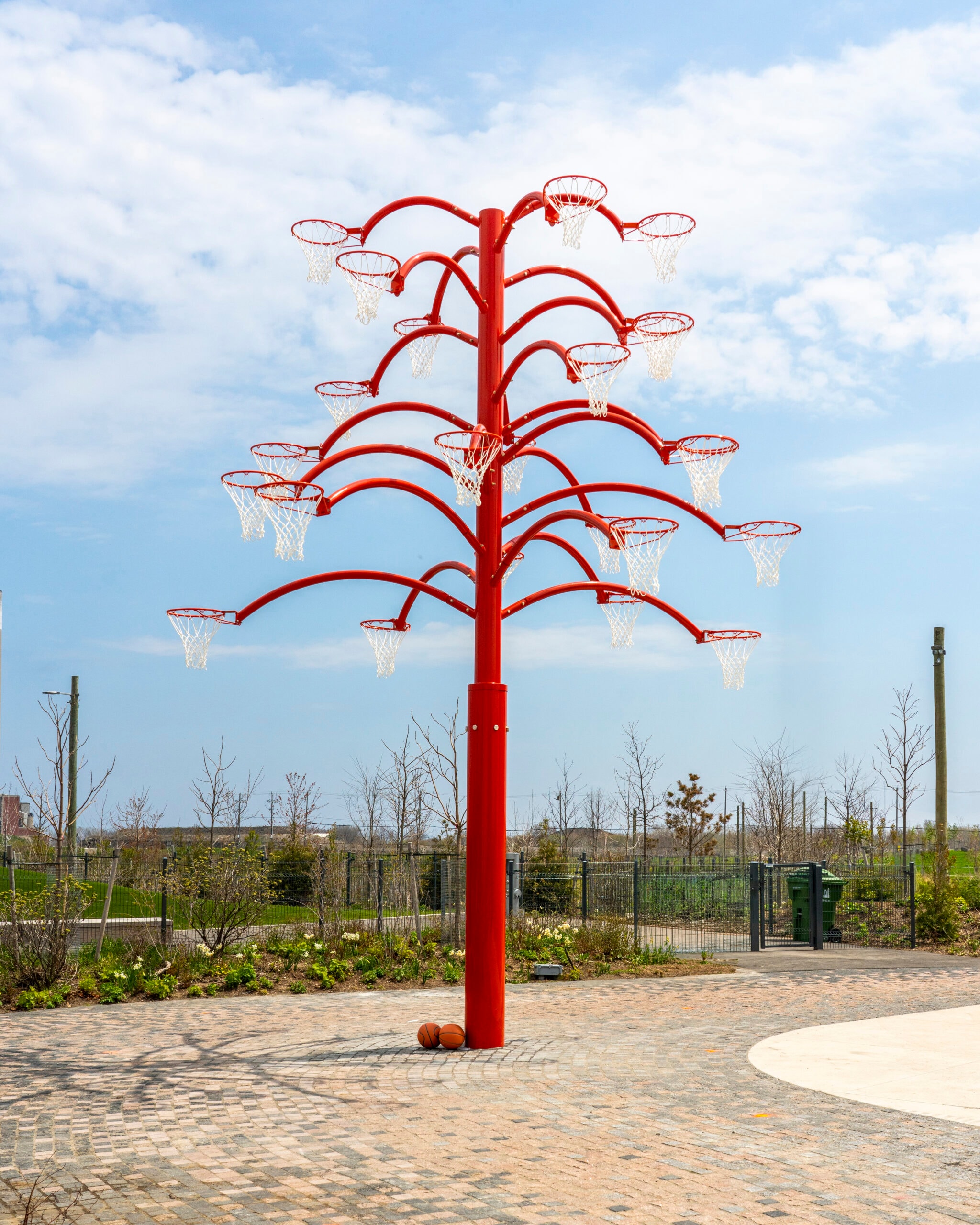A tall, bright red, tree-like sculpture with multiple basketball hoops on its branches stands on a brick-paved area. Two basketballs rest at its base.