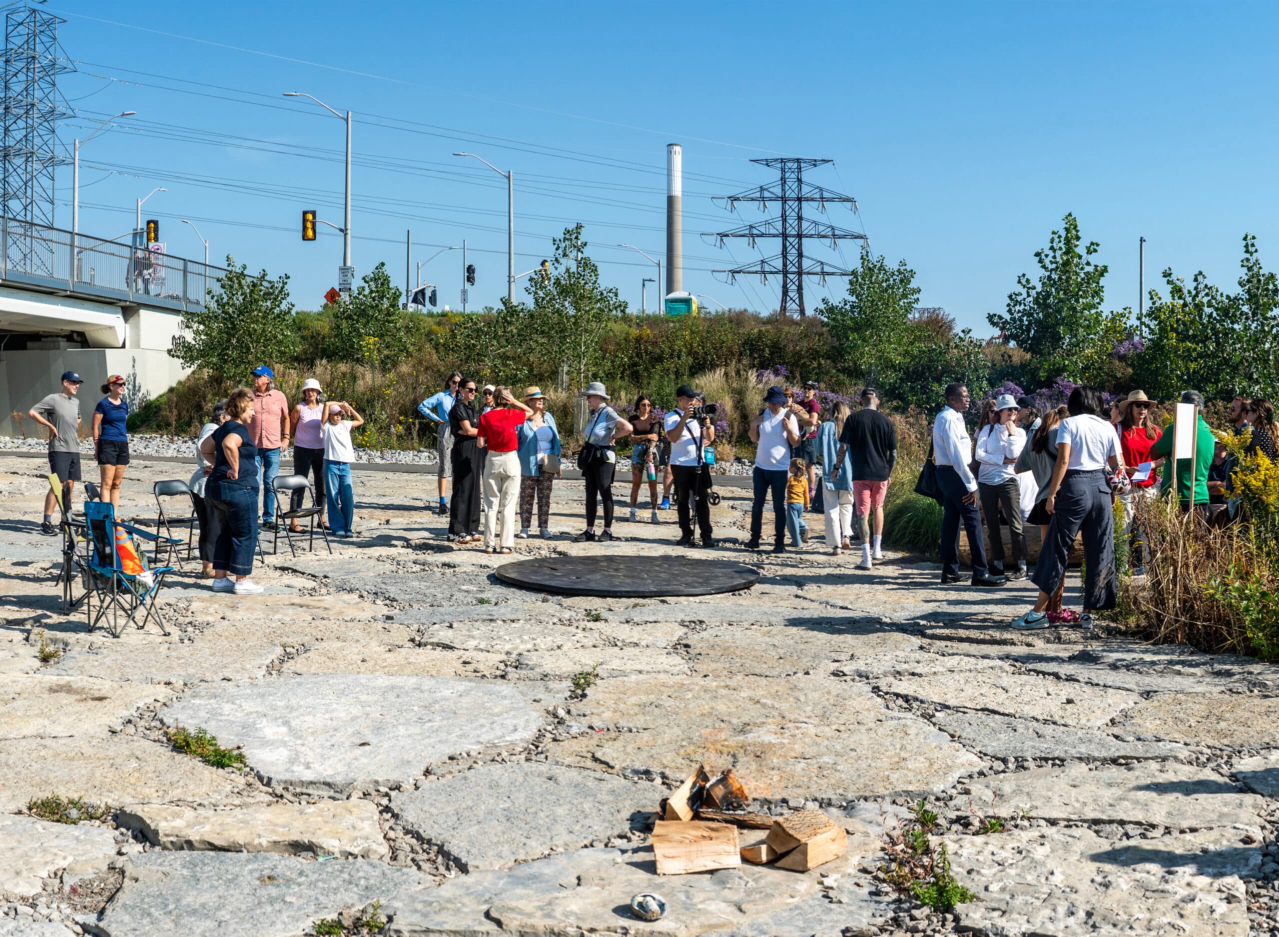 A group of people are gathered outdoors on a paved area with large stone slabs under a clear blue sky. In the background are power lines, a pylon, and a bridge. A bronze circular sculpture, Trajet by Caroline Monnet and Dean Baldwin Lew, is in the center foreground, with a small pile of wood nearby.