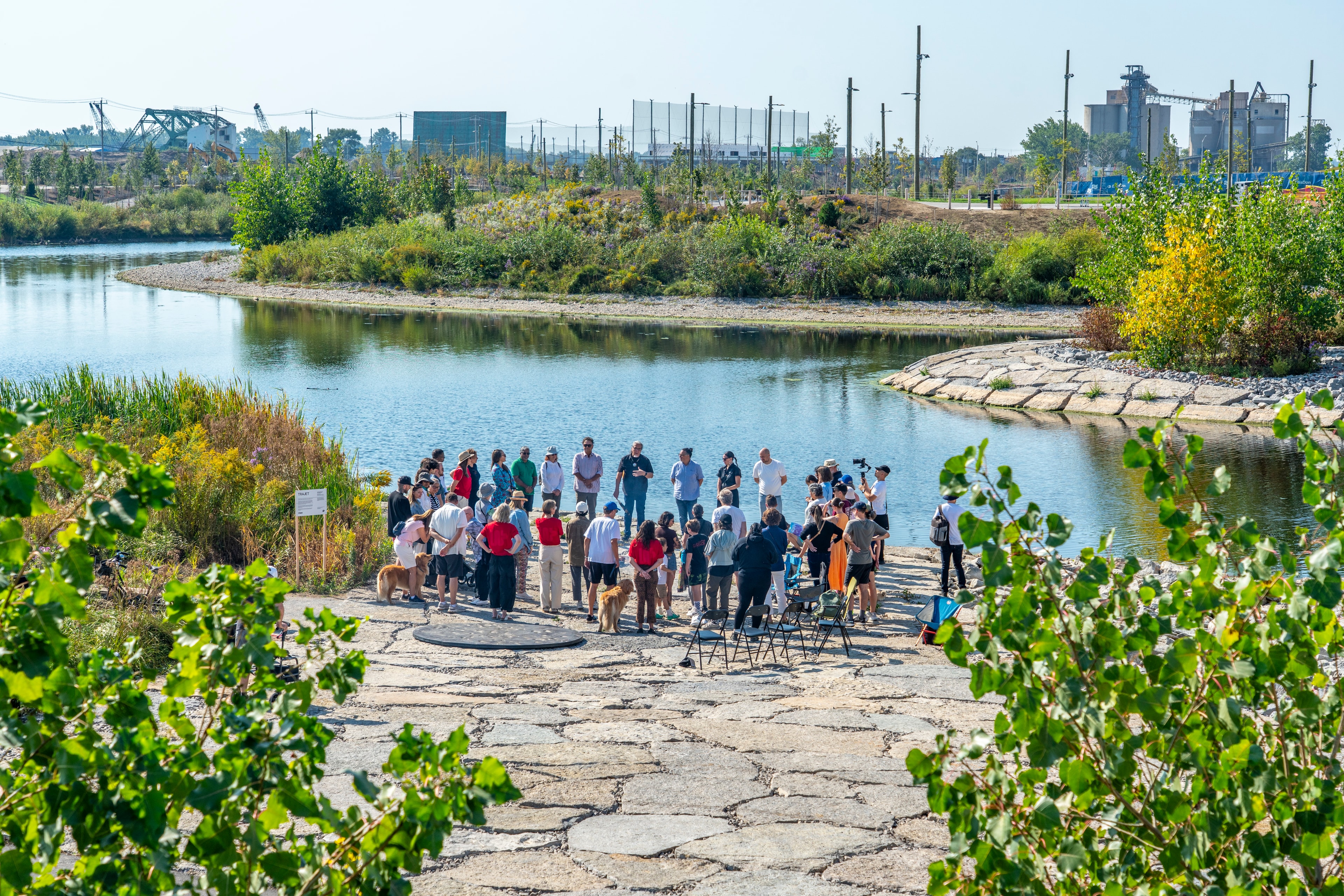 A group of people standing beside Trajet, a circular sculpture with foot imprints. Green and yellow foliage surrounds the outdoor area.