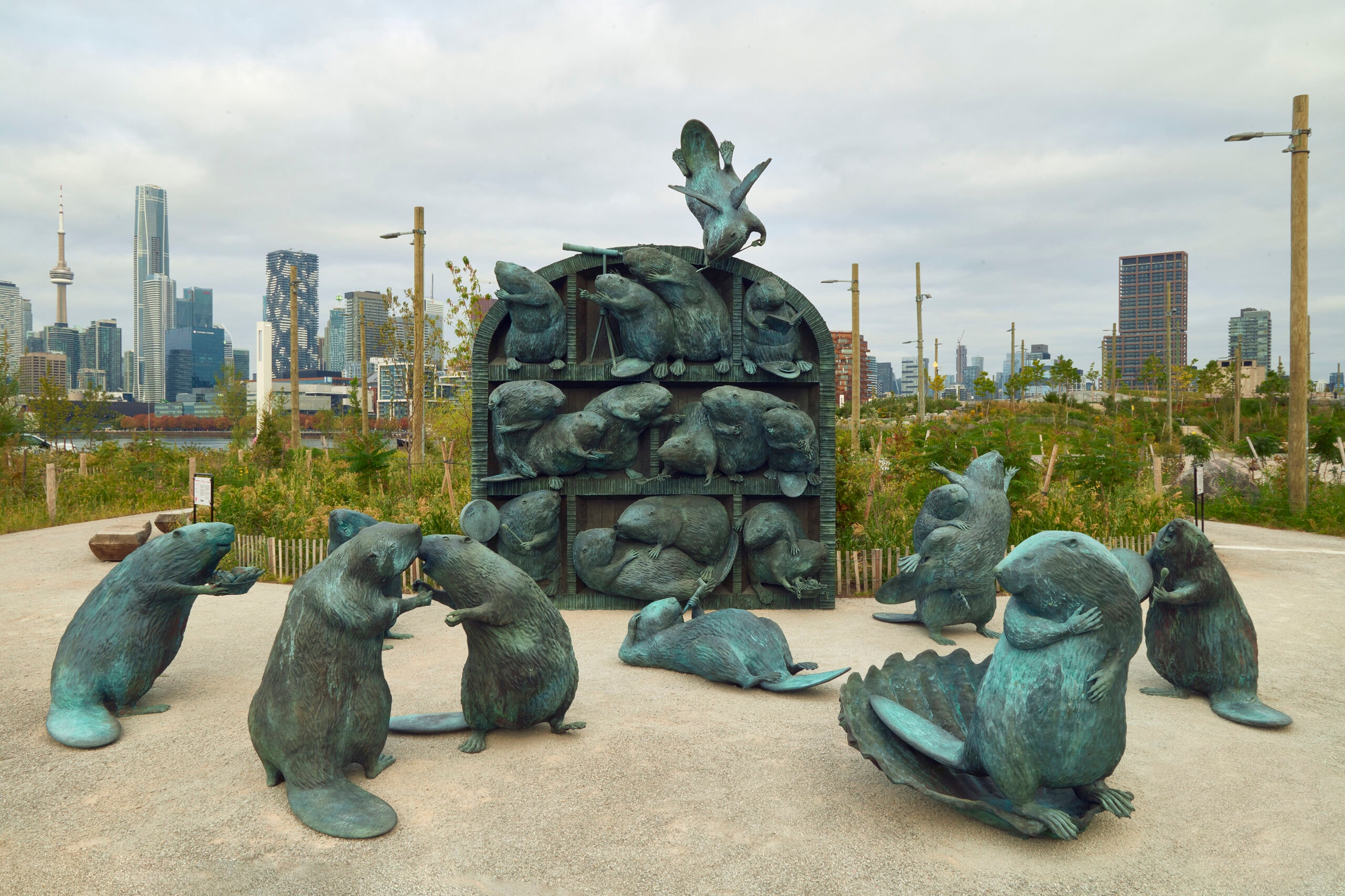 A large outdoor sculpture features numerous bronze beavers with a greenish patina. Some beavers are arranged within a dark, multi-shelved arched structure, while others are on the ground around it. A modern city skyline, including the CN Tower, is visible in the background under a cloudy sky