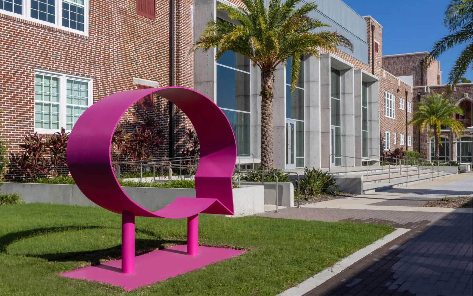 A large, bright pink sculpture shaped like a speech bubble. It rests on a flat base with two cylindrical supports, situated on a green lawn.