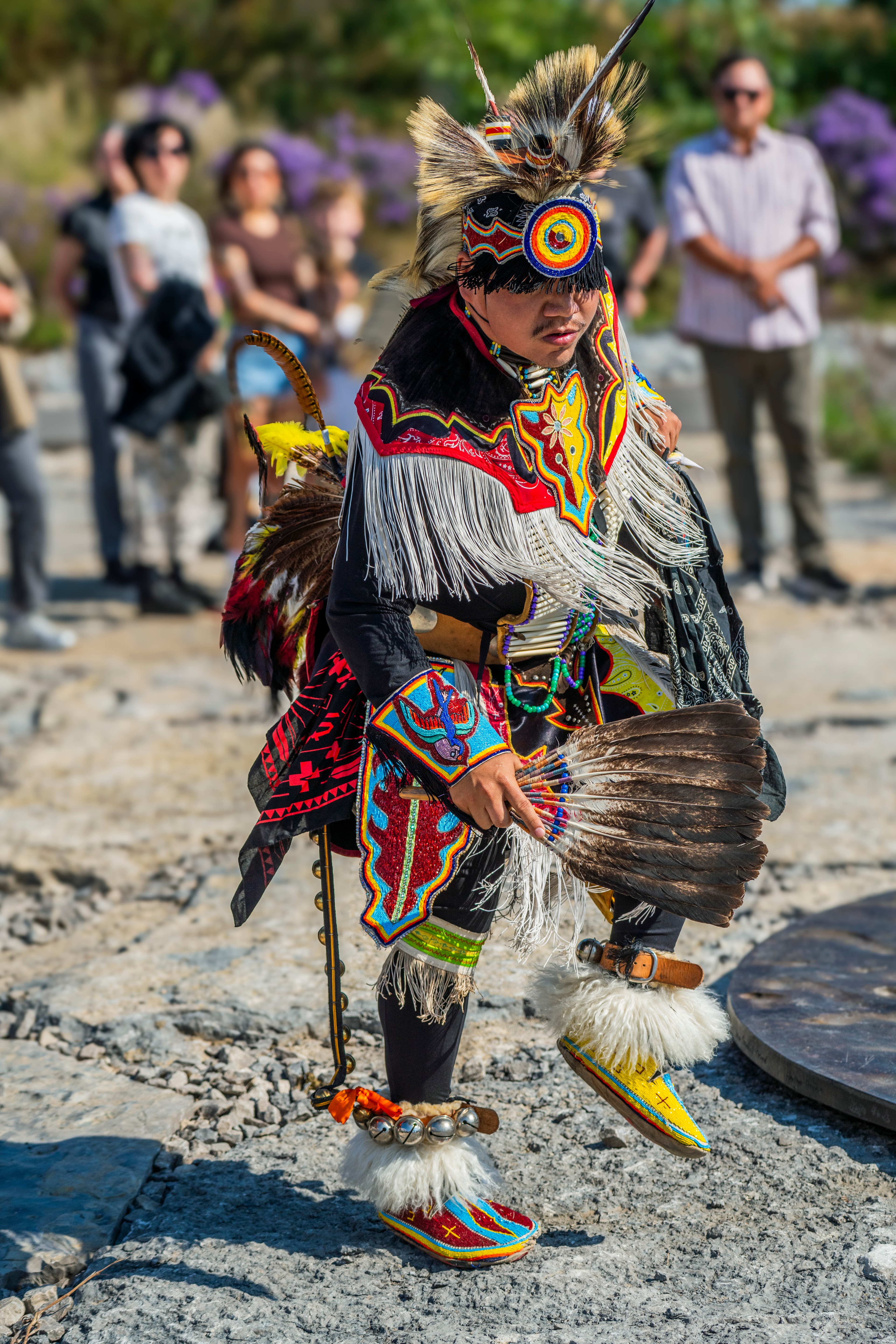 A person in vibrant traditional regalia, featuring a tall feathered headdress, colorful embroidered garments with white fringe, and beaded moccasins. They hold a large feather fan and wear bells around their ankles, standing on a textured outdoor surface.