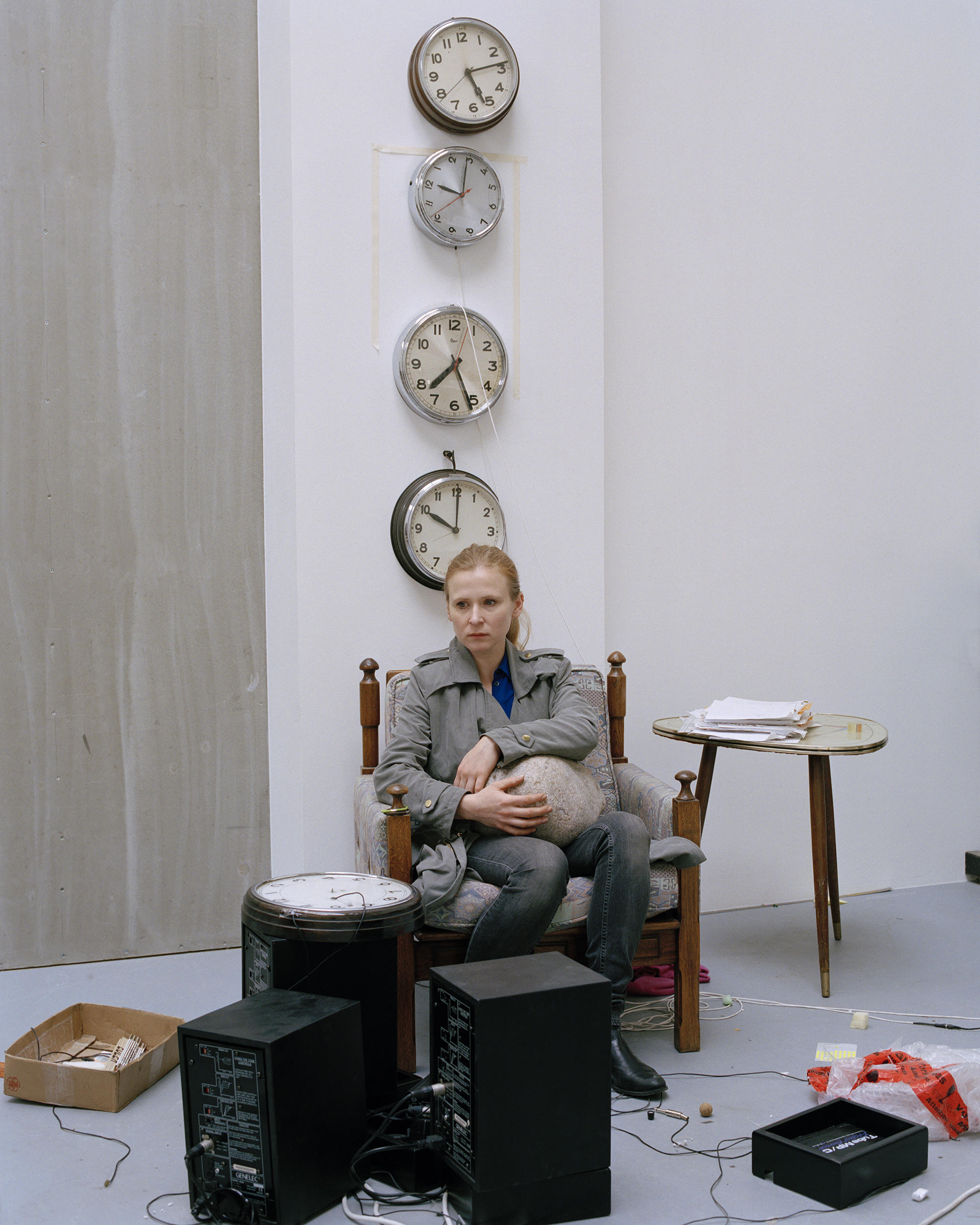 A woman with light hair sits in a patterned armchair, holding a spherical object. Behind her, four analog clocks are mounted vertically on a white wall. Various wires, speakers, and objects are scattered on the concrete floor around her.