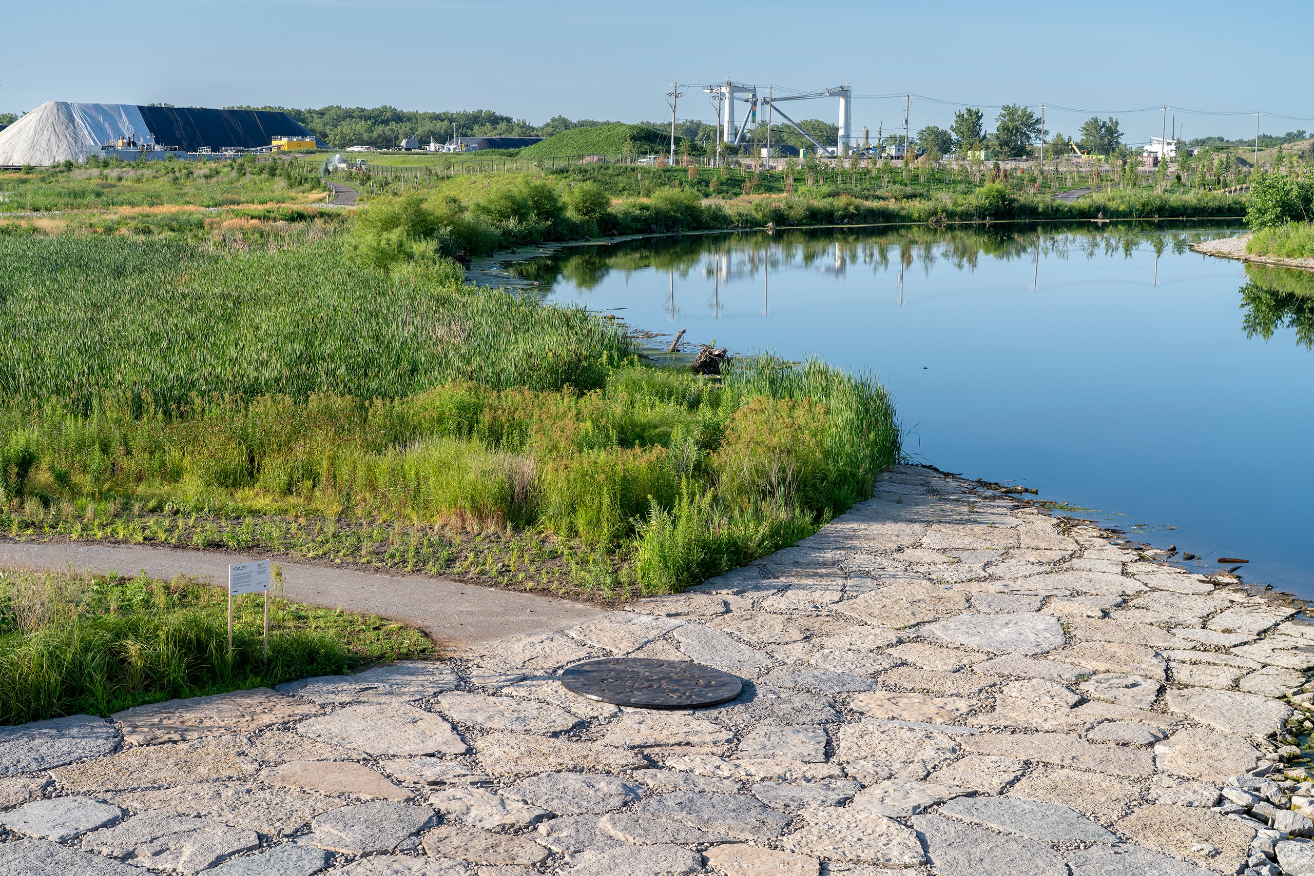 A large, dark circular sculpture with footprints embedded in its surface is integrated in a path of irregular stone slabs. The path curves through a grassy landscape with trees, and a tall industrial chimney stands in the distance.
